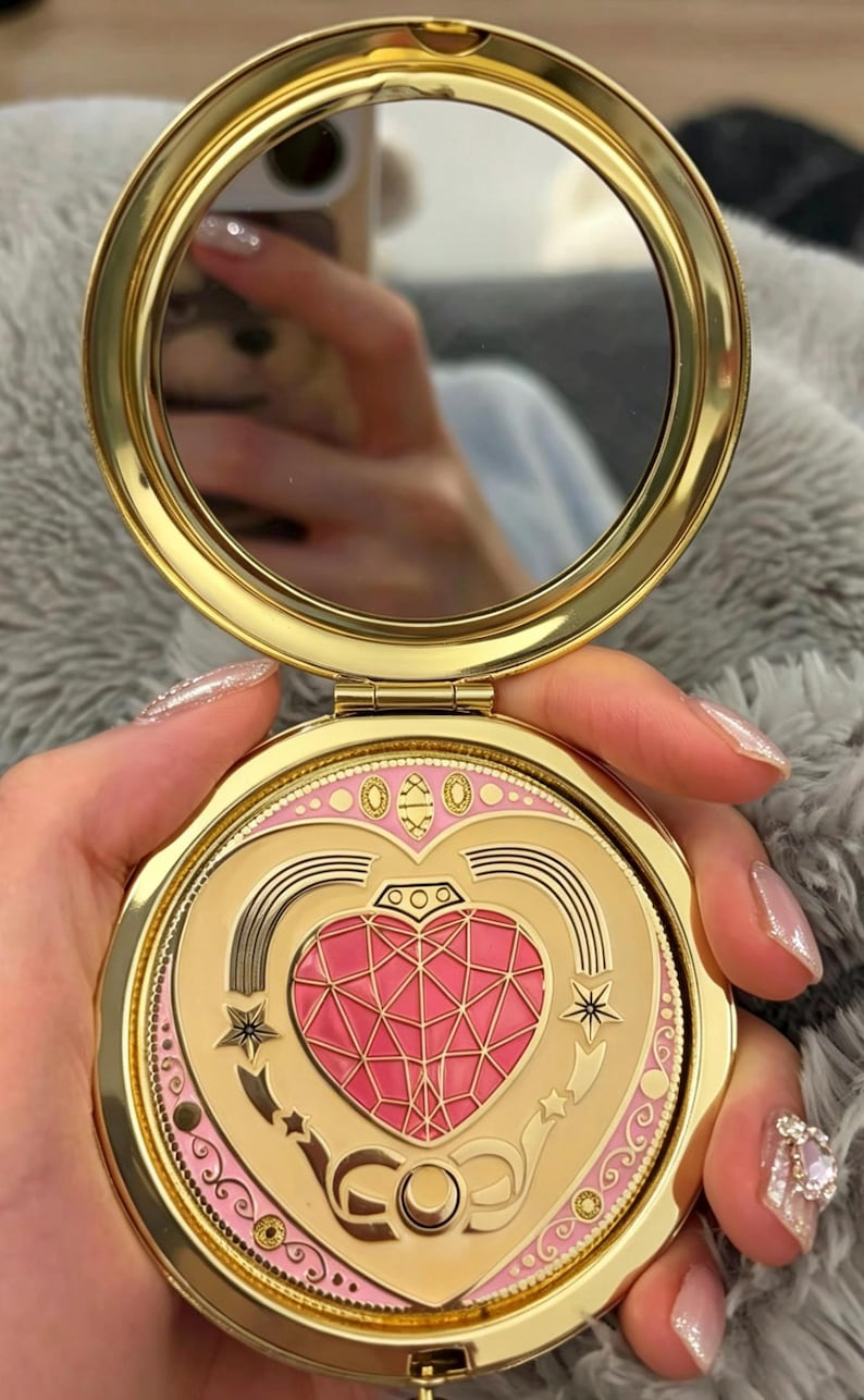 May include: A round gold-toned compact mirror featuring a hinged lid with a decorative design. The lid displays a central pink faceted heart emblem surrounded by gold star motifs, curved lines, and a pink border with small gold circular accents.