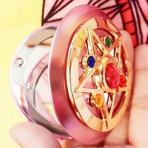 May include: A round pink compact mirror featuring a gold-toned star emblem on the lid. The star is adorned with small, colorful inset gems in red, blue, green, and yellow. The compact is open, revealing a reflective mirror surface inside the casing.