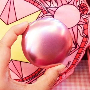 May include: A circular, metallic pink compact mirror with a smooth, reflective surface. The object is spherical in shape and features a solid, uniform rose-toned finish that catches the light, creating a subtle gradient across its curved exterior.