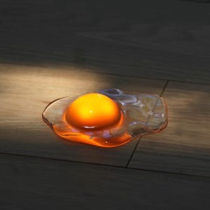 May include: A glowing, translucent, egg-shaped light fixture featuring a bright, rounded orange center that mimics a yolk, surrounded by a clear, irregular, flattened border that resembles egg white, emitting a warm, soft light.