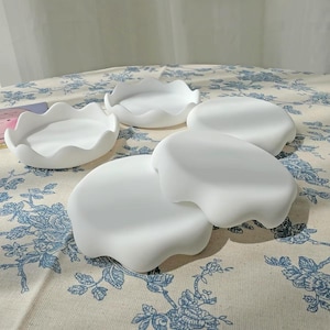 May include: A set of four white, circular trays with scalloped, wavy edges. Each tray features a smooth, matte finish and a shallow, concave centre. The items are arranged in a cluster, showcasing their uniform colour and distinct, curved rim design.