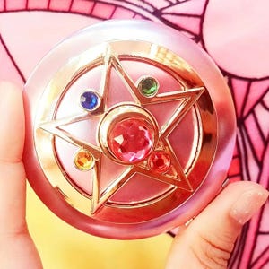 May include: A round, metallic pink compact case featuring a gold-toned star emblem on the lid. The star is adorned with a central faceted pink gem, surrounded by smaller blue, green, yellow, and red circular gems set within the gold star points.