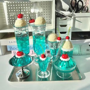 Könnte beinhalten: Eine Sammlung künstlicher blauer Gelatine-Desserts und Getränke in verschiedenen Glasbehältern, jeweils garniert mit einem Klecks weißer Sahne und einer leuchtend roten Kirsche. Das Set umfasst hohe Gläser, kleine Schalen und Schalen auf einem Silbertablett.