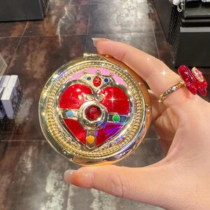 May include: A round gold compact mirror featuring a central red heart emblem adorned with small blue, green, yellow, and red gemstones. The design includes a crown-like top detail and a pink border, all set within a metallic gold frame.