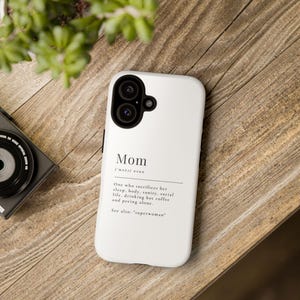 May include: White phone case featuring the word "Mom" and a definition printed in black. The definition includes the phrases "sacrifices her sleep, body, sanity, social life, drinking hot coffee and peeing alone." The case is on a wooden surface.