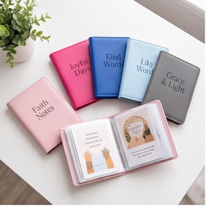 May include: A collection of five small rectangular journals in pink, magenta, blue, light blue, and gray, featuring the text Faith Notes, Joyful Days, Kind Words, Liky Word, and Grace & Light. One open pink journal displays two illustrated cards.
