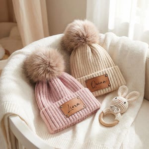 Pode incluir: Dois gorros de bebé de malha, um rosa e outro bege, cada um com um pompom de pele sintética e uma etiqueta de couro com um nome. Há também uma pequena chocalho de coelhinho de crochê. Os gorros estão sobre um cobertor branco.