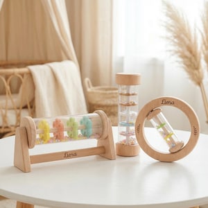 May include: Three wooden baby toys on a white table. The toys include a rolling rattle with colorful beads and shapes, a rain stick, and a circular rattle. The word "Liora" is engraved on two of the toys.
