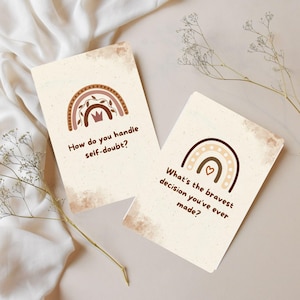 Puede incluir: Dos tarjetas beige con diseños de arcoíris y texto. Una tarjeta dice "¿Cómo manejas la duda?" y la otra dice "¿Cuál es la decisión más valiente que has tomado?" Las tarjetas tienen un efecto acuarela y elementos florales y de corona.