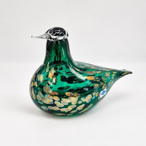 Op de afbeelding: Een glazen vogelbeeldje in een diepe smaragdgroene kleur, voorzien van een gespikkeld patroon met gouden en witte stippen over het hele lichaam. De kop is gemaakt van helder glas met zwarte accenten en een klein blauw-wit label is zichtbaar op de zijkant.