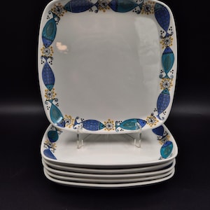 Può includere: Una pila di piatti quadrati in ceramica bianca. Ogni piatto presenta un bordo con pesci blu e verde acqua, con dettagli gialli e neri. I piatti sono impilati su un supporto trasparente su sfondo nero.