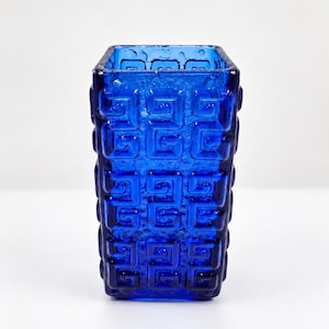 May include: A square-shaped cobalt blue glass vase featuring a repeating geometric Greek key pattern embossed on all four sides. The glass has a textured, slightly bubbly surface finish and a thick, open top rim with a deep blue translucent color.