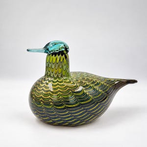 Op de afbeelding: Een glazen vogelbeeldje met een afgerond lichaam voorzien van een gelaagd, geschulpt patroon in tinten limoengroen, olijfgroen en donkerblauw. De kop is van effen doorschijnend lichtblauw met een bijpassende snavel en de staart loopt spits toe.