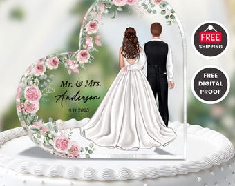 Könnte beinhalten: Ein herzförmiger Tortenaufsatz für eine Hochzeit mit einer Illustration von Braut und Bräutigam. Das Design umfasst rosa Rosen und Grün, mit den Namen "Mr. & Mrs. Anderson" und dem Datum "9.11.2025". Der Aufsatz befindet sich auf einer weißen Torte.