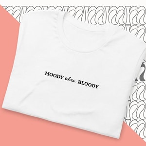 Puede incluir: Camiseta blanca con el texto negro "MOODY when BLOODY". La camiseta se muestra sobre un fondo rosa y crema con un patrón repetido. La camiseta tiene cuello redondo y mangas cortas.