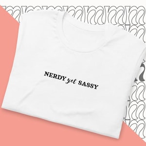 May include: A white t-shirt featuring the black text "NERDY yet SASSY". The t-shirt is made of a soft material and is folded on a pink surface with a decorative pattern in the background.