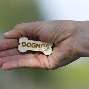 Può includere: Un'etichetta di legno a forma di osso di cane con la scritta "DOGNOS" incisa. L'etichetta presenta anche una piccola silhouette di cane. L'etichetta è tenuta in una mano aperta su uno sfondo verde sfocato.