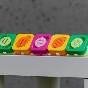 Może przedstawiać: Rząd pięciu kolorowych, kwadratowych przedmiotów z zaokrąglonymi wierzchami. Obiekty są zielone, żółte i różowe, każdy z kontrastującym okrągłym akcentem. Obiekty są ułożone na białej powierzchni na szarym tle.