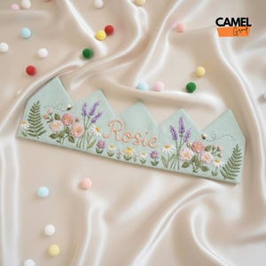 Può includere: Una corona di tessuto verde chiaro ricamata con fiori, felci e lavanda. Il nome "Rosie" è ricamato in corsivo. Una piccola ape è ricamata vicino ai fiori. La corona è su una superficie satinata.