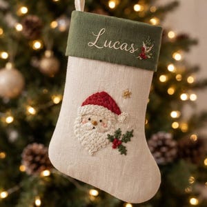 Op de afbeelding: Een beige kerstkous met een donkergroene boord waarop de naam Lucas en een klein bloemtakje zijn geborduurd. Het hoofdgedeelte toont een geborduurd gezicht van de Kerstman met een rode muts, versierd met groene hulstblaadjes en rode bessen.