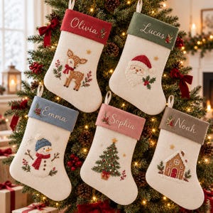 Op de afbeelding: Vijf crèmekleurige kerstsokken met geborduurde boorden in rood, groen, blauw, roze en grijs. Elke sok heeft een uniek kerstmotief: een reekalf, de Kerstman, een sneeuwman, een kerstboom en een peperkoekhuisje met namen erop geborduurd.