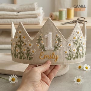 Puede incluir: Una corona de tela beige bordada con margaritas blancas y hojas verdes. El n&uacute;mero "1" y el nombre "Emily" est&aacute;n bordados en hilo blanco y dorado. La corona est&aacute; dise&ntilde;ada para una celebraci&oacute;n de primer cumplea&ntilde;os.