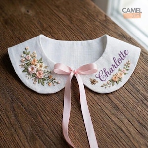 May include: White embroidered collar with floral and bee details. Features the name "Charlotte" in purple script. A pink ribbon is tied in a bow at the center. The collar is set against a wooden background.