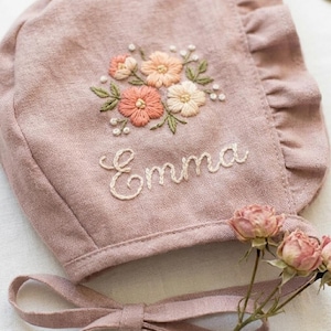 以下が含まれることがあります： ピーチ、クリーム、グリーンの色合いで刺繍された花のディテールとフリル付きの縁が特徴の、ダスティローズ色のリネン製ボンネット。生地の中央には白い糸の筆記体でEmmaという名前が刺繍されており、小さなバラが添えられています。