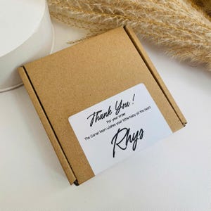 Peut inclure: Une petite bo&icirc;te en carton marron avec une &eacute;tiquette blanche. L'&eacute;tiquette indique "Thank You!" en cursive, avec du texte suppl&eacute;mentaire en dessous. Le nom "Rhys" est &eacute;crit en cursive. La bo&icirc;te est sur une surface blanche.