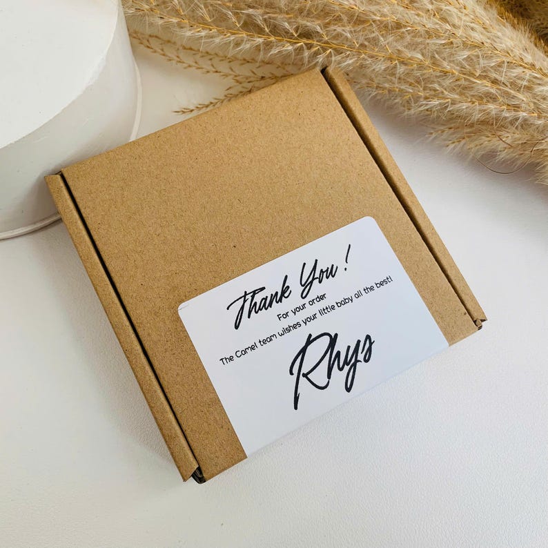 Peut inclure: Une petite bo&icirc;te en carton marron avec une &eacute;tiquette blanche. L'&eacute;tiquette indique "Thank You!" en cursive, avec du texte suppl&eacute;mentaire en dessous. Le nom "Rhys" est &eacute;crit en cursive. La bo&icirc;te est sur une surface blanche.