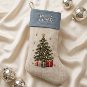 Könnte beinhalten: Ein beigefarbener Weihnachtsstrumpf aus Leinen mit einem blauen Umschlag, der mit dem Wort Noel bestickt ist. Der Körper zeigt einen gestickten grünen Weihnachtsbaum mit roten und weißen Kugeln, einem goldenen Stern und drei Geschenken.