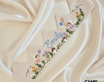 Könnte beinhalten: Ein beiges Stoffkrone mit gestickten Details. Die Krone zeigt ein Feen-Design mit bunten Luftballons, Blumen und dem Namen "Emily" in blauer Schrift. Eine Biene ist ebenfalls auf der Krone gestickt. Die Krone ist für ein Kind bestimmt.