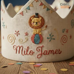 Peut inclure: Couronne en lin brod&eacute; sur le th&egrave;me du cirque. Elle pr&eacute;sente un lion en &eacute;quilibre sur une balle, des accents color&eacute;s et le nom "Milo James" en &eacute;criture rouge. D&eacute;cor&eacute;e de petits drapeaux et d'&eacute;toiles.