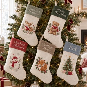 Könnte beinhalten: Fünf Weihnachtsstrümpfe in Leinenoptik mit bestickten Bündchen und festlichen Motiven. Die Bündchen tragen die Texte: Baby's First Christmas, Merry Christmas, Our First Christmas, Christmas Magic und Noel. Motive: Rehkitz, Bär, Schneemann, Fuchs und Weihnachtsbaum.