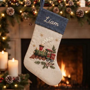 Pode incluir: Uma meia de Natal de linho bege com um punho azul-escuro bordado com o nome Liam em letra branca. O corpo da meia apresenta uma locomotiva a vapor bordada em verde e vermelho com fumo branco e ramos de pinheiro decorativos.