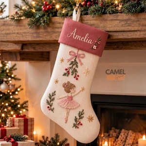 May include: A cream-coloured Christmas stocking featuring a dusty rose cuff embroidered with the name Amelia and floral accents. The body displays an embroidered ballerina in a pink tutu, a mistletoe sprig with a bow, and gold star motifs.