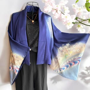 以下が含まれることがあります： マネキンに展示された、袖に花柄模様のあるネイビーブルーの着物風ジャケット。 ジャケットは黒いトップスとパンツと組み合わせています。 幾何学的なペンダントが付いたネックレスが見えます。