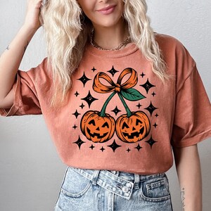 May include: A terracotta-colored short-sleeve t-shirt featuring a graphic print of two orange jack-o'-lantern pumpkins shaped like cherries. The pumpkins are connected by green stems with a large orange bow on top, surrounded by black star icons.