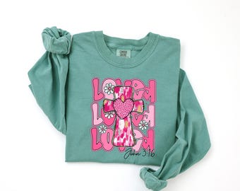 May include: A teal long-sleeve shirt with a graphic design. The design features the word "LOVED" stacked three times, with a pink cross and heart in the center. White daisies and the text "John 3:16" are also included.