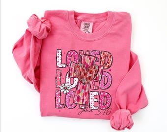 May include: A pink long-sleeved shirt with the word "LOVED" in a decorative font, featuring a cross and heart design. The text "John 3:16" is printed at the bottom. The shirt is made of a soft material.