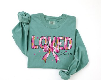 May include: A teal long-sleeve shirt with the word "LOVED" in pink and yellow block letters. A pink bow with yellow accents is below the word. The text "John 3:16" is printed below the bow.