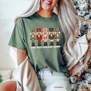 May include: An olive green short-sleeve t-shirt featuring a graphic of five nutcracker figures in various colors and patterns, with the text "THE NUTCRACKER CREW" printed in white capital letters underneath the figures on the front of the shirt.