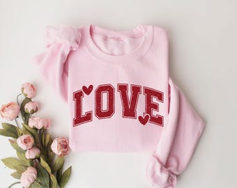 May include: A light pink sweatshirt with the word "LOVE" in a distressed red font. The letter "O" is replaced with a heart, and there are two additional hearts. The sweatshirt is folded, and a bouquet of pink flowers is in the corner.
