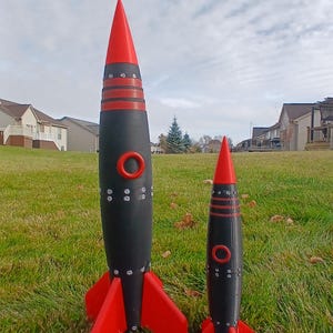 May include: Two toy rockets with a retro design. The rockets have a black body with red fins, a red nose cone, and a red ring around the middle. The rockets are standing on a grassy lawn.