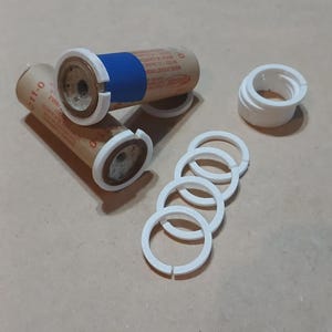 May include: Model rocket components, including brown cardboard tubes with white and blue sections, white plastic rings, and a white plastic cylinder. The parts are arranged on a light brown surface.