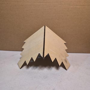 May include: A light-coloured wooden structure shaped like a stylised evergreen tree, featuring a pointed peak and jagged, stepped edges. The two sides meet at a central vertical seam, creating a tent-like form with a smooth, natural wood grain finish.