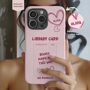 May include: A pink phone case with a library card design. The case features the name "Olivia" in a heart, the words "BOOKS MADE ME THIS WAY" and "NO REGRETS". The case is MagSafe compatible.