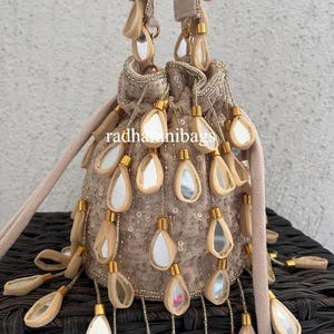 以下が含まれることがあります： ベージュ色の巾着バッグ。ミラーアクセントとビーズタッセル付き。バッグはスパンコールボディと柔らかい明るい色のストラップが特徴です。「radharanibags」の文字がバッグに表示されています。