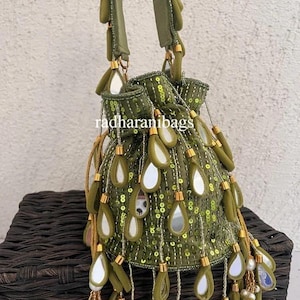 May include: Olive green beaded drawstring bag with teardrop-shaped embellishments and mirror accents. The bag features a drawstring closure, gold-toned accents, and the text "radharanibags". The bag is sitting on a woven basket.