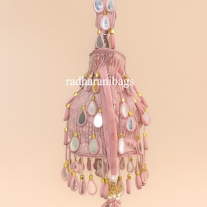 May include: A pink, bell-shaped bag with decorative mirror accents and dangling teardrop-shaped embellishments. The bag has a long handle with similar embellishments and the text "radharanibags".